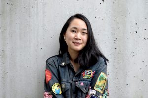 Headshot of a woman wearing a denim jacket covered in patches, leaning against a concrete wall