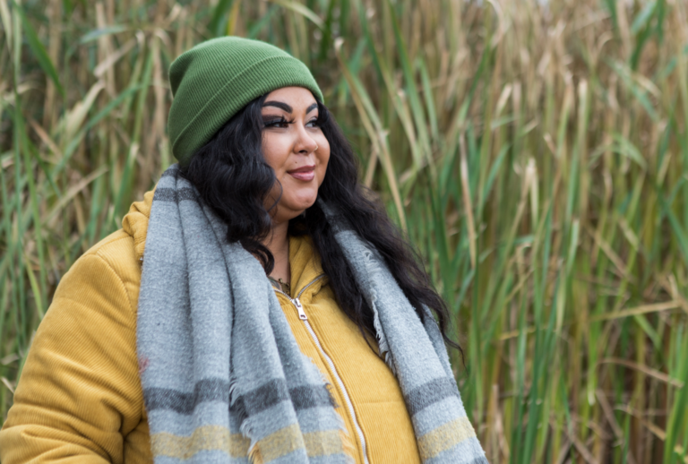 A woman with dark brown hair stands in a field, wearing a green toque, yellow jacket, and gray scarf.