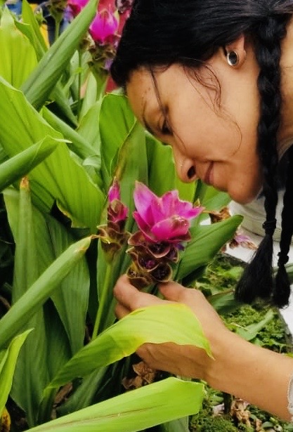 Carolynne leans down to smell a pink flower. She has silver circular earrings, and two braids in her dark brown hair. 