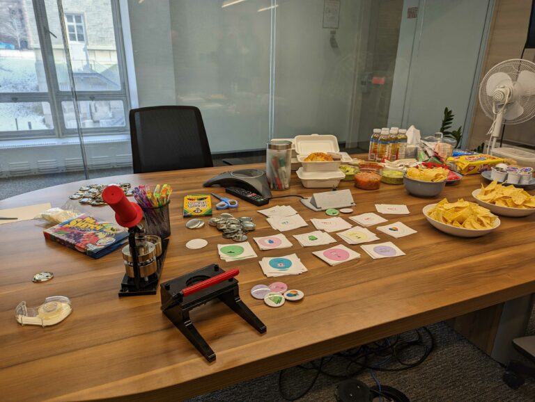 A boardroom table covered in chips, salsa, and other snacks. There is also a button maker and supplies to make buttons and pins with fun designs.