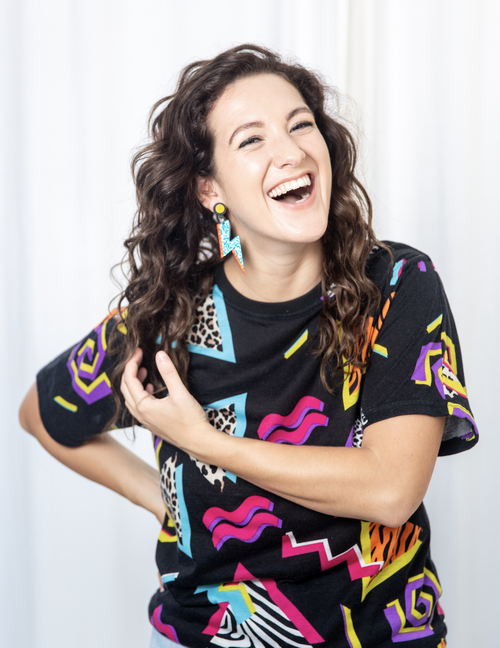 A woman with long curly hair wearing teal lightening bolt earrings laughs at the camera. She is wearing a black shirt with 80s-style designs in blue, pink, and yellow.