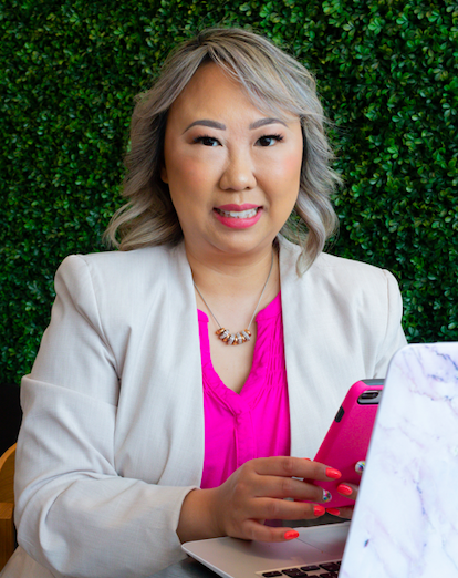 A headshot of Vera Cheng. She is wearing a white blazer and a bright pink shirt, and sitting in front of a plant-covered wall. She has bright pink nails and it is holding a pink cellphone, in front of a laptop. She has silver hair and is smiling.