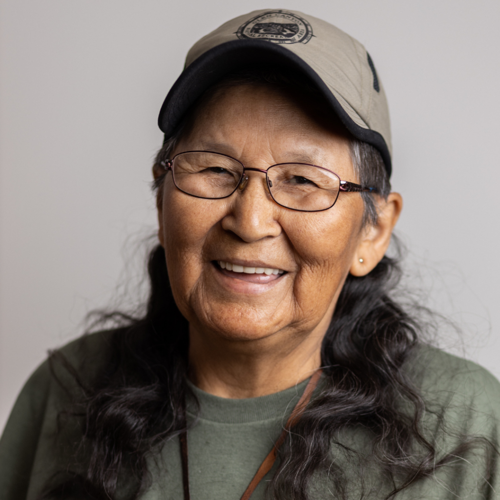 Ma-Nee Chacaby smiles at the camera. She is wearing a gray baseball cap, wire-framed glasses, and a green t-shirt.
